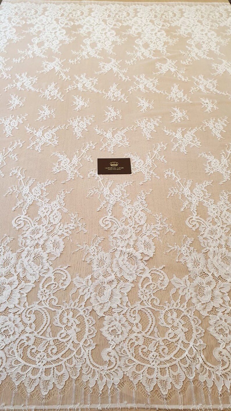 May include: A white lace fabric featuring an intricate floral pattern with scalloped edges and a sheer mesh base. A small rectangular black tag with a gold crown icon and the text IMPERIAL LACE is placed in the center of the fabric.