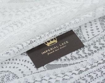 May include: White lace fabric with a delicate floral pattern. The fabric is draped over a brown card with the text "Imperial Lace French Lace Dealer".