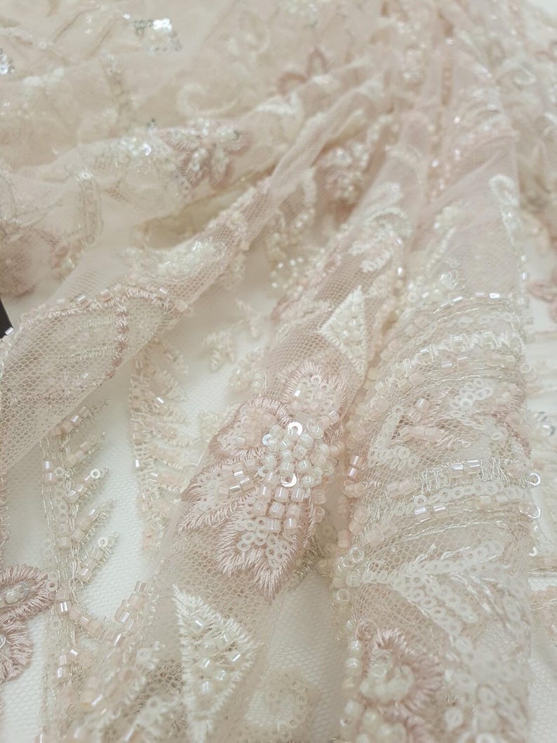 May include: Close-up of delicate, beaded lace fabric in a blush pink and ivory colour scheme. The lace features intricate floral patterns and small, iridescent beads.