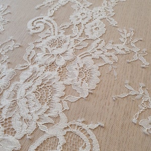 May include: White floral lace fabric featuring intricate embroidered blossoms and leafy vines layered over a solid beige mesh base. The delicate needlework creates a textured pattern with scalloped edges and openwork details throughout the textile.