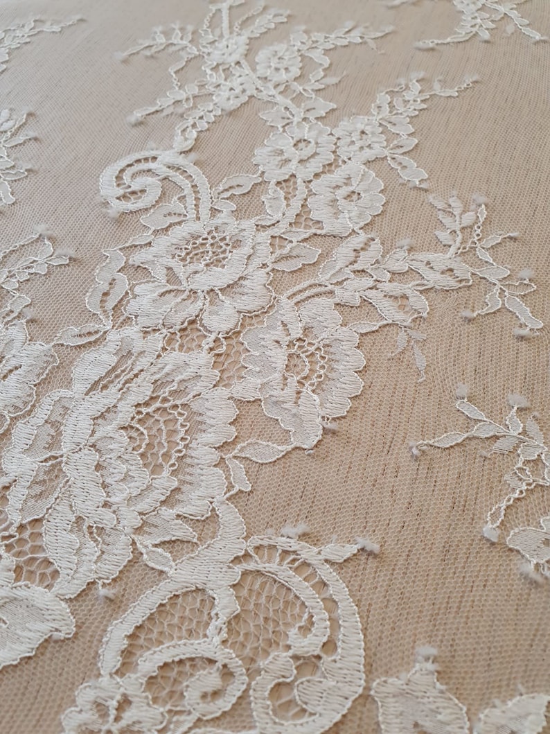 May include: White floral lace fabric featuring intricate embroidered blossoms and leafy vines layered over a solid beige mesh base. The delicate needlework creates a textured pattern with scalloped edges and openwork details throughout the textile.
