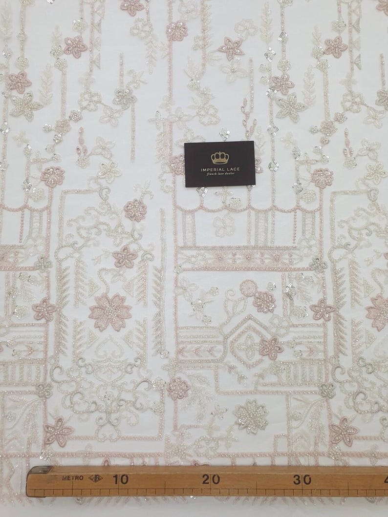 May include: White beaded lace fabric with a geometric pattern and pink floral accents. The fabric is shown on a white background with a ruler measuring 76 cm.
