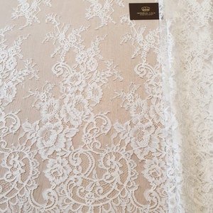 May include: White lace fabric featuring an intricate floral pattern with scalloped edges. A small dark rectangular tag is visible on the fabric with the text Imperial Lace French lace dealer and a crown icon printed in gold lettering.