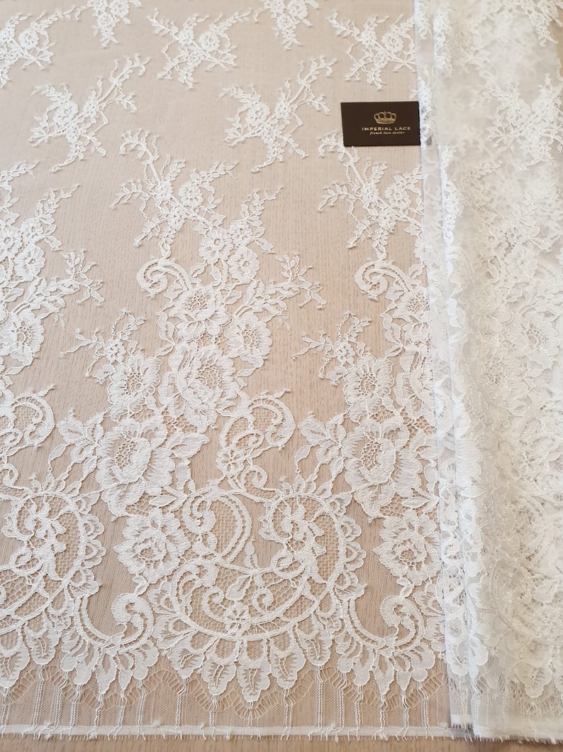 May include: White lace fabric featuring an intricate floral pattern with scalloped edges. A small dark rectangular tag is visible on the fabric with the text Imperial Lace French lace dealer and a crown icon printed in gold lettering.