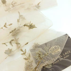 Puede incluir: Primer plano de encaje francés color marfil con bordado floral dorado con cuentas. La delicada tela está doblada, mostrando los intrincados detalles del diseño. Una etiqueta con el texto "IMPERIAL LACE french lace dealer" es visible.