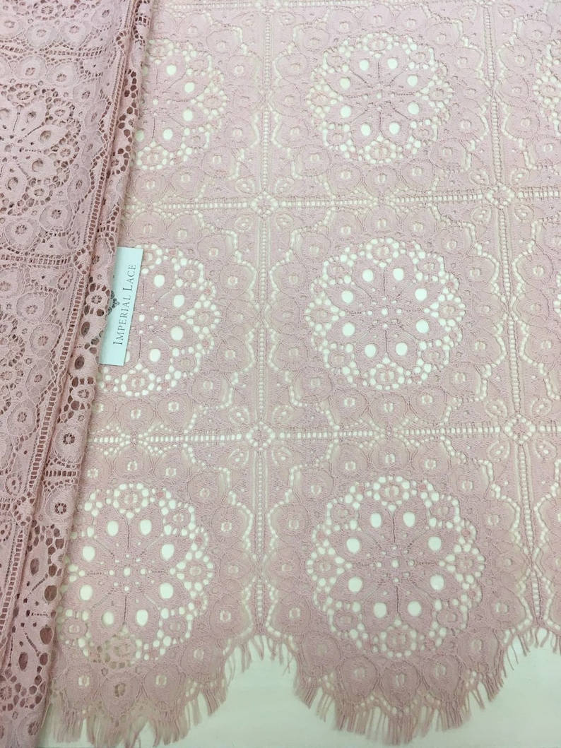 May include: Pink lace fabric with a delicate floral pattern and scalloped edges. The fabric has a square grid pattern with a circular design in each square. The label reads "Imperial Lace".