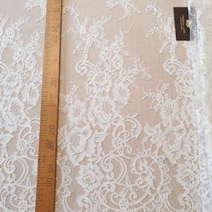 May include: White lace fabric featuring an intricate floral pattern with scalloped edges. A wooden ruler is placed vertically along the left side, showing measurements in centimeters up to 60 cm. A small black tag reads IMPERIAL LACE.