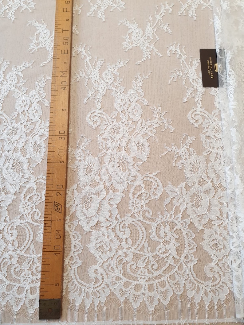 May include: White lace fabric featuring an intricate floral pattern with scalloped edges. A wooden ruler is placed vertically along the left side, showing measurements in centimeters up to 60 cm. A small black tag reads IMPERIAL LACE.