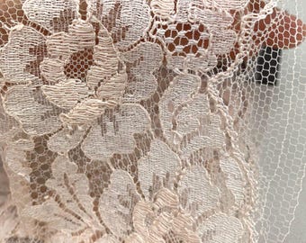 May include: Close-up of delicate, pale pink lace fabric. The lace features a floral pattern with detailed, raised designs. The fabric has a fine, net-like background, and the overall texture appears soft and intricate. The lace is likely used for apparel or craft projects.