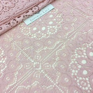 May include: Pink lace fabric with a delicate floral pattern. The fabric has a soft, flowing texture and is perfect for creating garments, accessories, or home decor. The label reads "Imperial Lace".