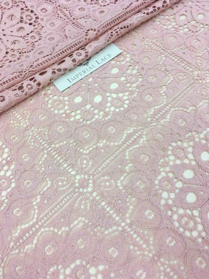 May include: Pink lace fabric with a delicate floral pattern. The fabric has a soft, flowing texture and is perfect for creating garments, accessories, or home decor. The label reads "Imperial Lace".