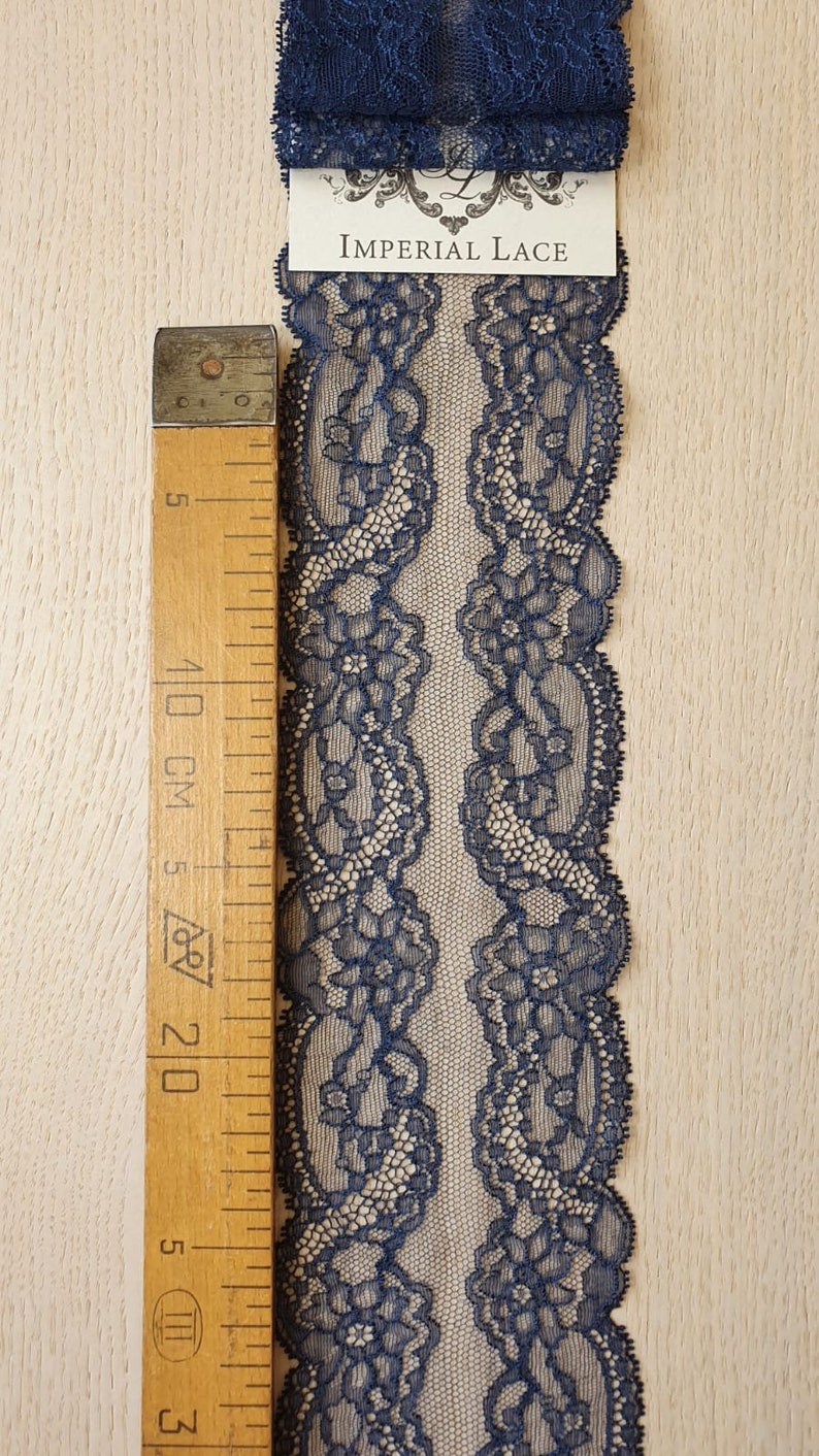 May include: A close-up of navy blue lace trim with a floral pattern. The lace is measured against a wooden ruler with markings in inches and centimeters. The label reads "Imperial Lace".
