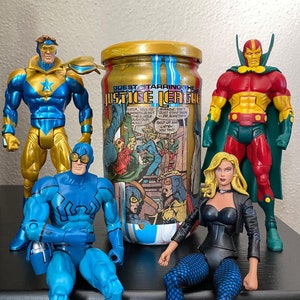 May include: A collection of Justice League action figures, including Booster Gold, Blue Beetle, and others, are arranged around a jar with a gold lid and the words "GUEST STARRING THE JUSTICE LEAGUE" printed on it. The jar has comic book art on it.