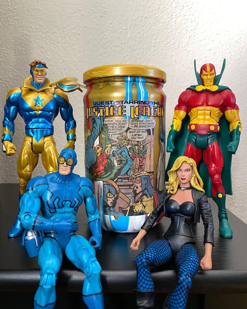 May include: A collection of Justice League action figures, including Booster Gold, Blue Beetle, and others, are arranged around a jar with a gold lid and the words "GUEST STARRING THE JUSTICE LEAGUE" printed on it. The jar has comic book art on it.