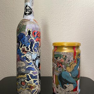 May include: A tall wine bottle and a small jar, both decorated with comic book panels. The bottle has a DC logo on top and the words "CAPTAIN ATOM" on the side. The jar has a gold lid and comic book art.