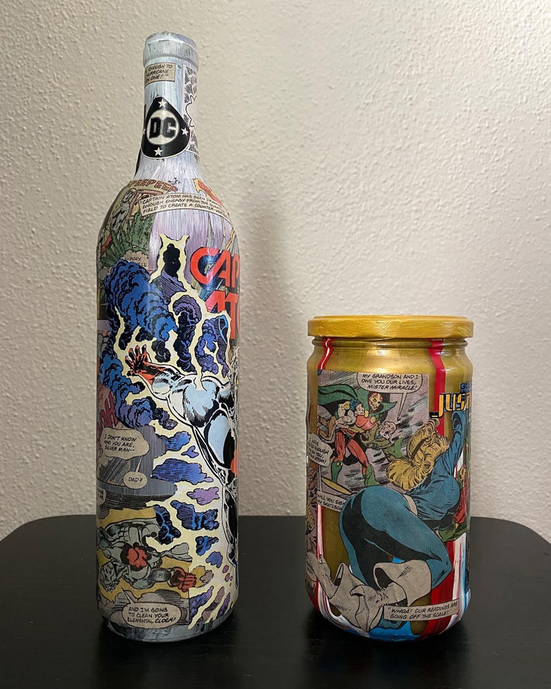 May include: A tall wine bottle and a small jar, both decorated with comic book panels. The bottle has a DC logo on top and the words "CAPTAIN ATOM" on the side. The jar has a gold lid and comic book art.