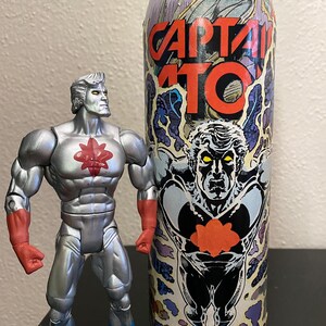 May include: A silver action figure with red accents and blue boots stands next to a tall, cylindrical container. The container features the words "CAPTAIN ATOM" in red, with a comic book-style illustration.