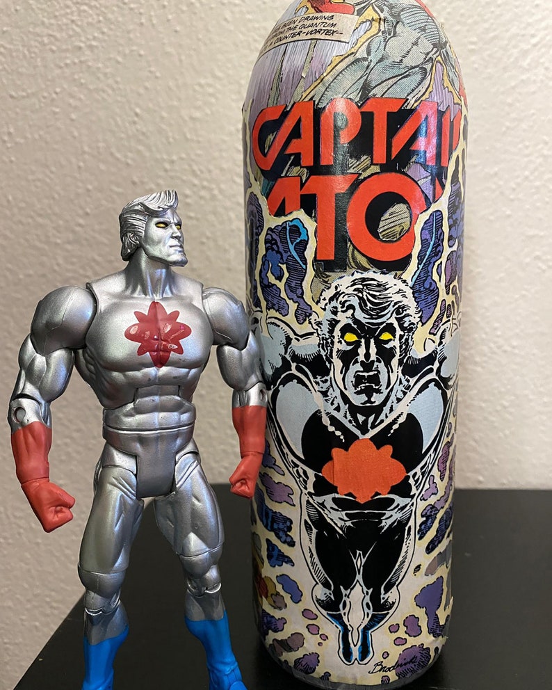 May include: A silver action figure with red accents and blue boots stands next to a tall, cylindrical container. The container features the words "CAPTAIN ATOM" in red, with a comic book-style illustration.