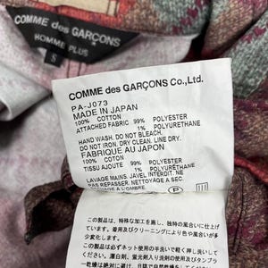 May include: A close-up of a label from a Comme des Garcons Homme Plus garment. The white label has black text detailing the garment's composition: 100% cotton and 99% polyester. Washing instructions are included, along with the text "MADE IN JAPAN."