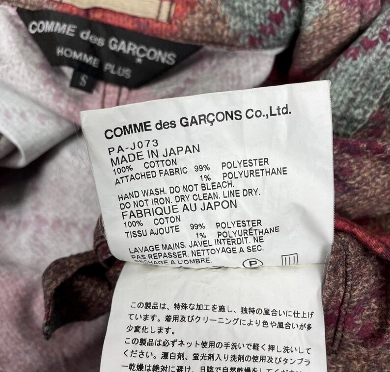 May include: A close-up of a label from a Comme des Garcons Homme Plus garment. The white label has black text detailing the garment's composition: 100% cotton and 99% polyester. Washing instructions are included, along with the text "MADE IN JAPAN."