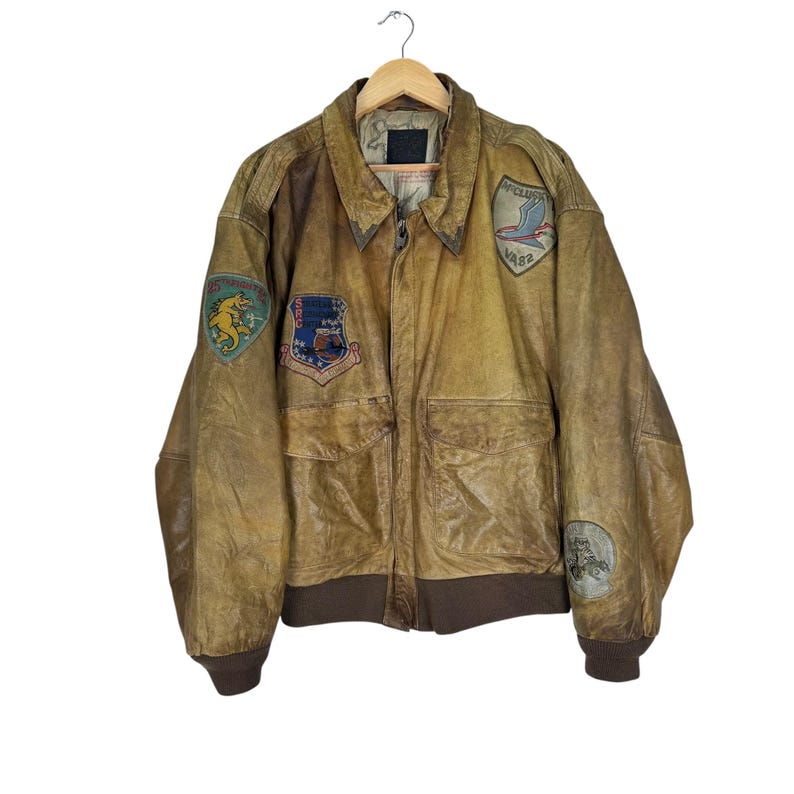 May include: A vintage brown leather bomber jacket with multiple patches. The jacket has a collared neckline, two front pockets, and ribbed cuffs and hem. Patches include a shield with stars and a winged emblem.