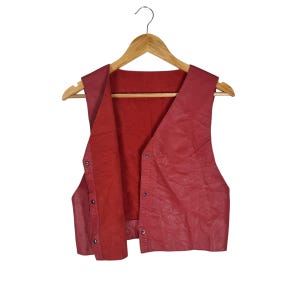 Op de afbeelding: Een rode leren vest met een V-hals hangt aan een houten hanger. Het vest heeft een knoopsluiting aan de voorkant en een getextureerde oppervlakte. De binnenvoering is ook rood.