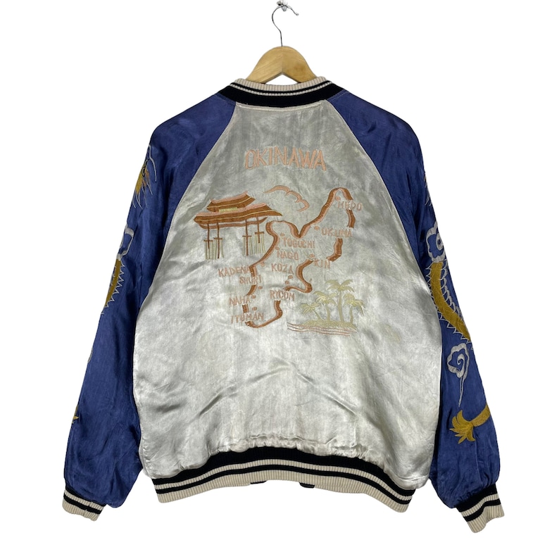 May include: A blue and silver satin bomber jacket with an embroidered map of Okinawa, Japan on the back. The map includes the names of various cities and towns on the island. The jacket has a black and white striped collar and cuffs.