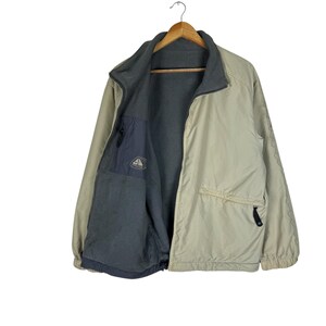 Puede incluir: Chaqueta con cremallera beige y gris con forro polar gris. La chaqueta tiene cuello alto, bolsillo en el pecho y dos bolsillos laterales. La chaqueta est&aacute; colgada en una percha de madera.