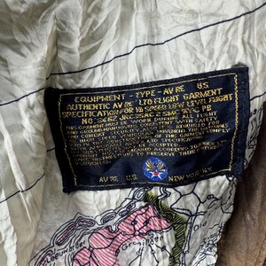 May include: Close-up of a vintage brown leather jacket interior. The lining is a cream-colored fabric with a map design. A black label with gold lettering details the jacket's specifications, including "AUTHENTIC AV RE LTD FLIGHT GARMENT."