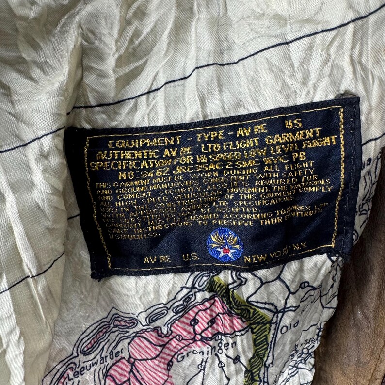 May include: Close-up of a vintage brown leather jacket interior. The lining is a cream-colored fabric with a map design. A black label with gold lettering details the jacket's specifications, including "AUTHENTIC AV RE LTD FLIGHT GARMENT."