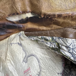May include: Close-up of a distressed brown leather jacket, revealing a cream-colored lining with a map design. The leather shows wear and tear, with visible cracks and discoloration, suggesting age and use.