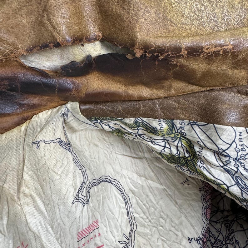 May include: Close-up of a distressed brown leather jacket, revealing a cream-colored lining with a map design. The leather shows wear and tear, with visible cracks and discoloration, suggesting age and use.