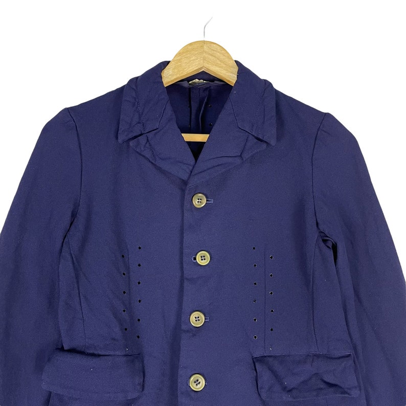 Puede incluir: Una chaqueta azul marino con cuello y cinco botones. La chaqueta tiene dos bolsillos de solapa y un patr&oacute;n decorativo de peque&ntilde;os agujeros en la parte delantera.