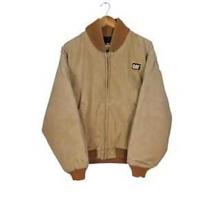 May include: A tan-colored bomber jacket with a brown ribbed collar, cuffs, and hem. The jacket has a zipper closure and two front pockets. A small patch with the word "CAT" is on the upper right chest.