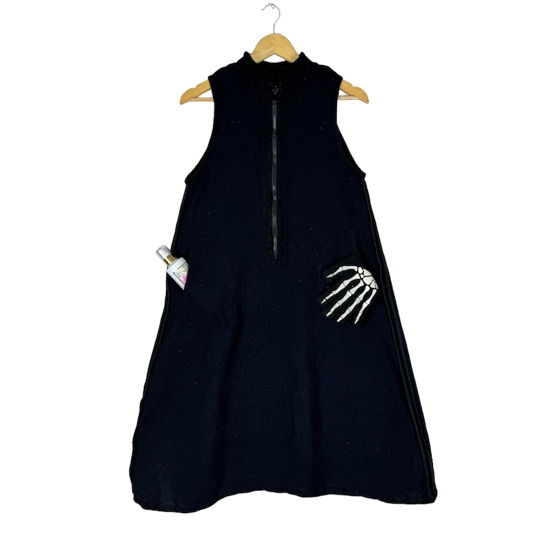 K&ouml;nnte beinhalten: Ein schwarzes &auml;rmelloses Kleid mit einem Rei&szlig;verschluss an der Vorderseite und einem Skelett-Hand-Design auf der rechten Seite. Das Kleid hat eine Tasche auf der linken Seite.