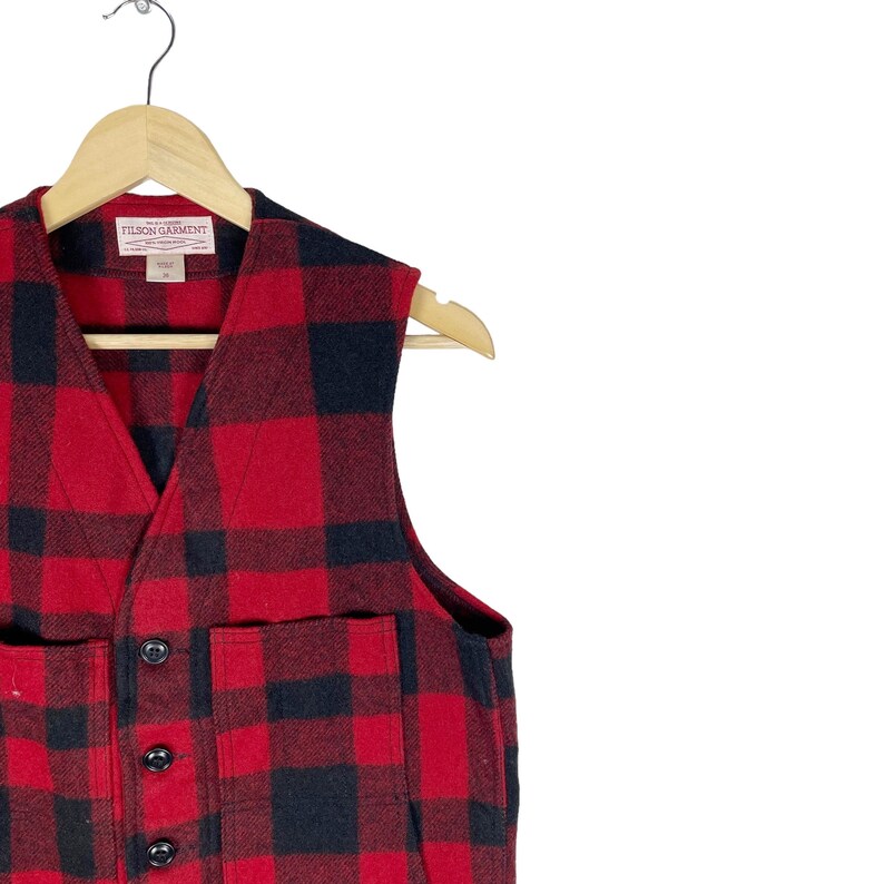 Op de afbeelding: Een rood en zwart geruit vest met knoopsluiting en twee zakken. Het vest heeft een Filson Garment-label aan de voorkant.
