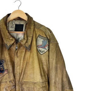 May include: A vintage brown leather jacket with a worn, distressed appearance. The jacket features a collared neckline, a zipper closure, and multiple patches, including one with a bird emblem and the text "McClusky VA82". The jacket is hanging on a wooden hanger.