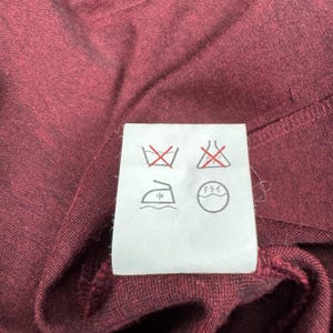 Op de afbeelding: Close-up van een bordeauxrode stof met een wit waslabel. Het label toont verschillende wassymbolen, waaronder een doorgestreept wassymbool, een symbool voor niet bleken, een strijksymbool en een stomerijsymbool.