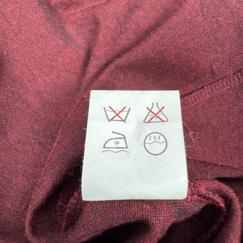Op de afbeelding: Close-up van een bordeauxrode stof met een wit waslabel. Het label toont verschillende wassymbolen, waaronder een doorgestreept wassymbool, een symbool voor niet bleken, een strijksymbool en een stomerijsymbool.