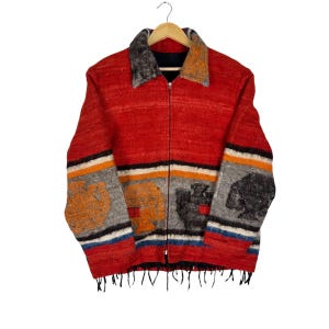 May include: A red zip-up jacket with a textured weave. The jacket features a collar and cuffs with a gray and orange pattern. The sleeves have colorful stripes and patterned designs in orange, gray, and black. The jacket has a fringe at the bottom.