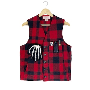 Op de afbeelding: Een rood en zwart geruit flanellen vest met een skelet hand patch op de linkerzak en een zwarte zak aan de rechterkant. Het vest heeft een knoopsluiting en hangt aan een houten hanger.