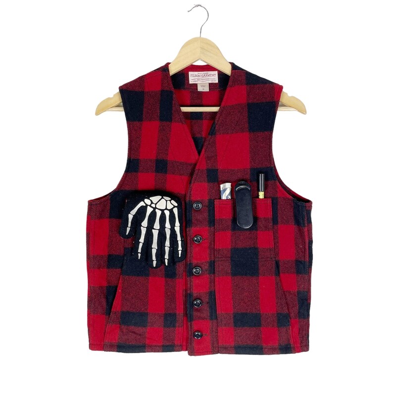 Op de afbeelding: Een rood en zwart geruit flanellen vest met een skelet hand patch op de linkerzak en een zwarte zak aan de rechterkant. Het vest heeft een knoopsluiting en hangt aan een houten hanger.