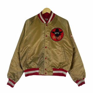 Op de afbeelding: Een gouden satijnen varsity jack met rode en witte geribbelde boorden. Het jack heeft een grote geborduurde patch op de linker borst met de tekst "Outlaws" met een gemaskerde cowboy met een hoed. Het jack heeft een knoopsluiting en twee zakken.