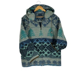 May include: A hooded jacket with a full zipper, featuring a pattern of snowflakes, trees, and geometric shapes in shades of blue, teal, and gray. The jacket is displayed on a wooden hanger.