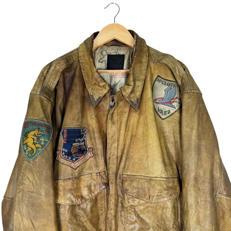 May include: A vintage brown leather bomber jacket with multiple patches. The jacket has a zipper closure, two front pockets, and a collar. Patches include a dragon, a flying bird, and text reading "25th Fighter Sq" and "VA82".