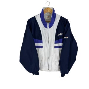 May include: A vintage navy blue, white, and purple track jacket with a full zipper. The jacket features the brand name "ellesse" on the chest and sleeve. The jacket is hanging on a wooden hanger.