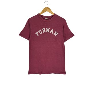 May include: A maroon short-sleeved t-shirt with a crew neckline, featuring the word FURMAN printed in a white arched font across the chest. The shirt is displayed on a wooden hanger, showcasing a textured fabric finish and a standard fit.