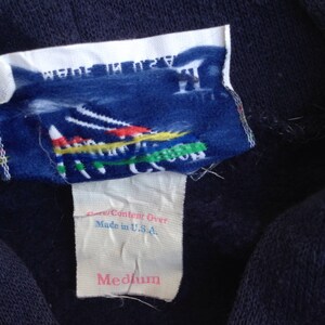 K&ouml;nnte beinhalten: Ein marineblauer Sweatshirt mit einem wei&szlig;en Etikett, auf dem "Fiber Content Over Made in U.S.A. Medium" steht. Das Etikett zeigt au&szlig;erdem eine bunte Grafik einer Bergkette.