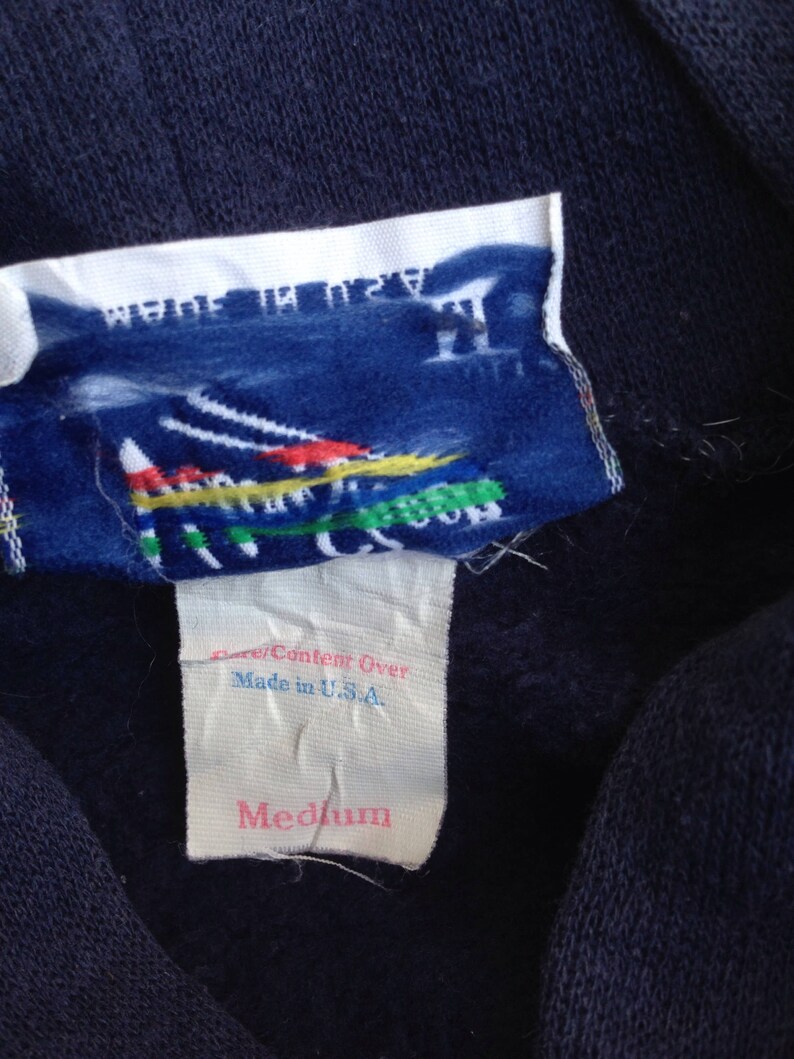 K&ouml;nnte beinhalten: Ein marineblauer Sweatshirt mit einem wei&szlig;en Etikett, auf dem "Fiber Content Over Made in U.S.A. Medium" steht. Das Etikett zeigt au&szlig;erdem eine bunte Grafik einer Bergkette.