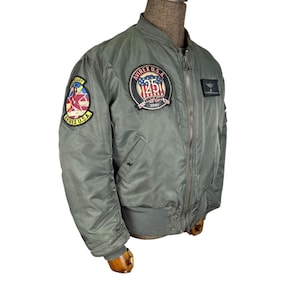 Peut inclure: Veste de bombardier vert olive avec plusieurs écussons, dont un écusson du 25e anniversaire. La veste a un col, des poignets et un ourlet côtelés. Fermeture à glissière et deux poches avant. La veste est présentée sur un mannequin.